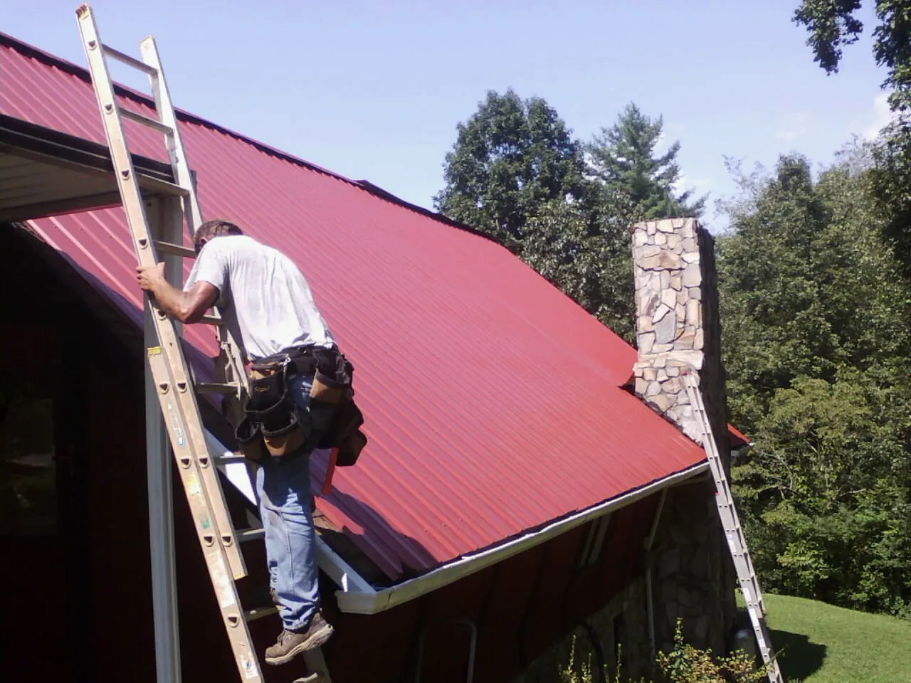 Roofer on a ladder beside a red metal roof and stone chimney — Soffit & Fascia Repair by Roofing Brothers in Reidsville